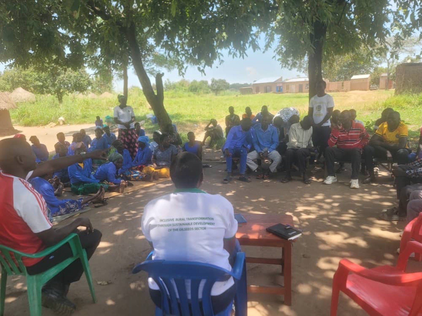 A farmer group meeting under trees in Nwoya District, with a National Oil Seeds Project facilitator addressing members