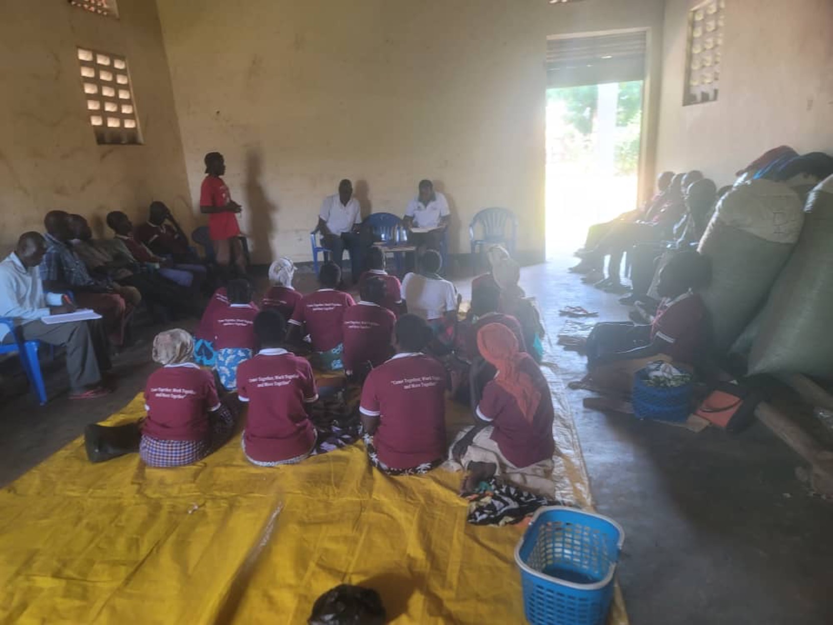 Members of a farmer group meeting inside a store, seated on tarpaulins in matching maroon group shirts with produce sacks stacked behind them